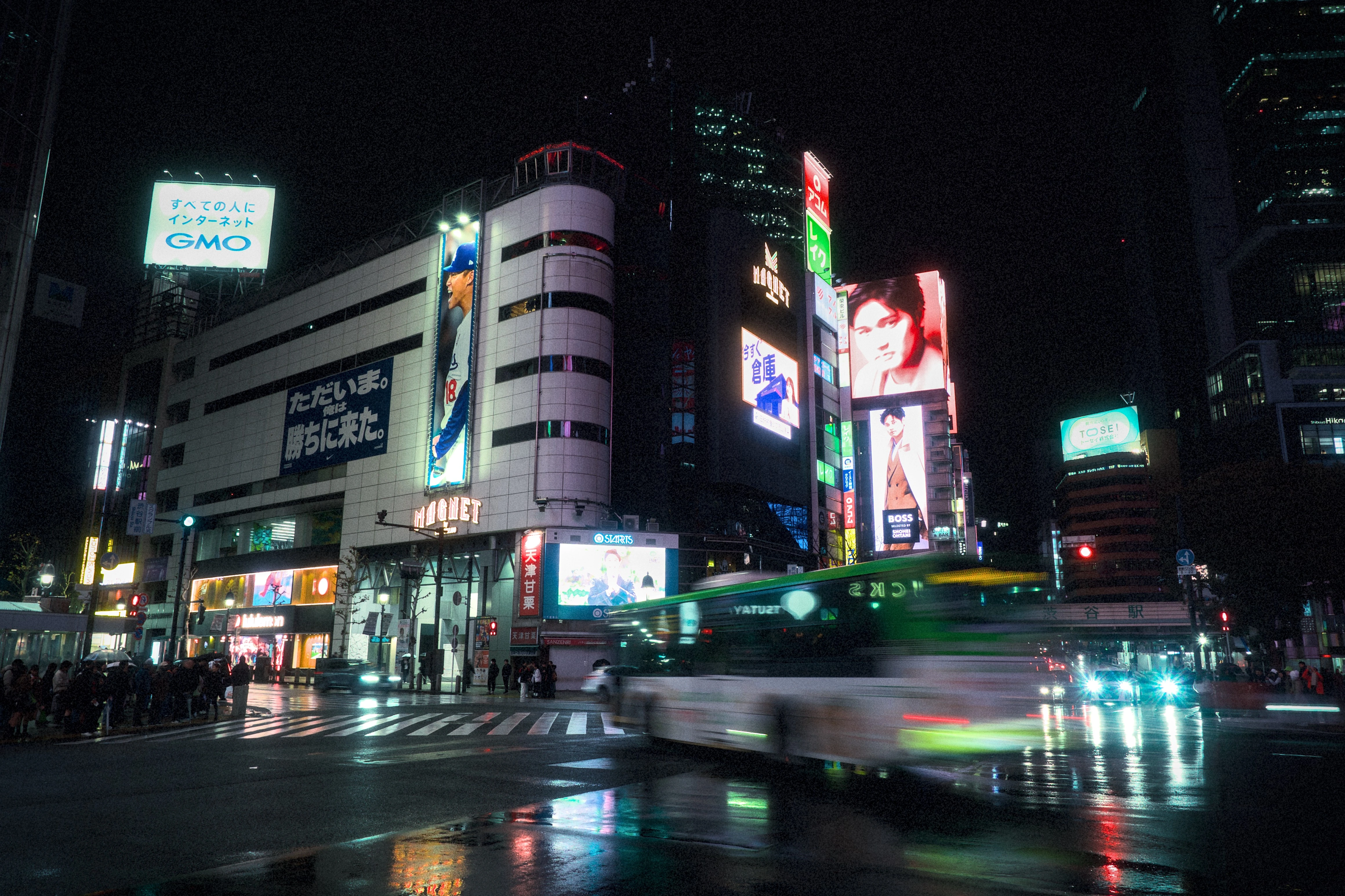 Night street scene with wet reflections and motion blur from traffic, shot handheld at a slow shutter speed.