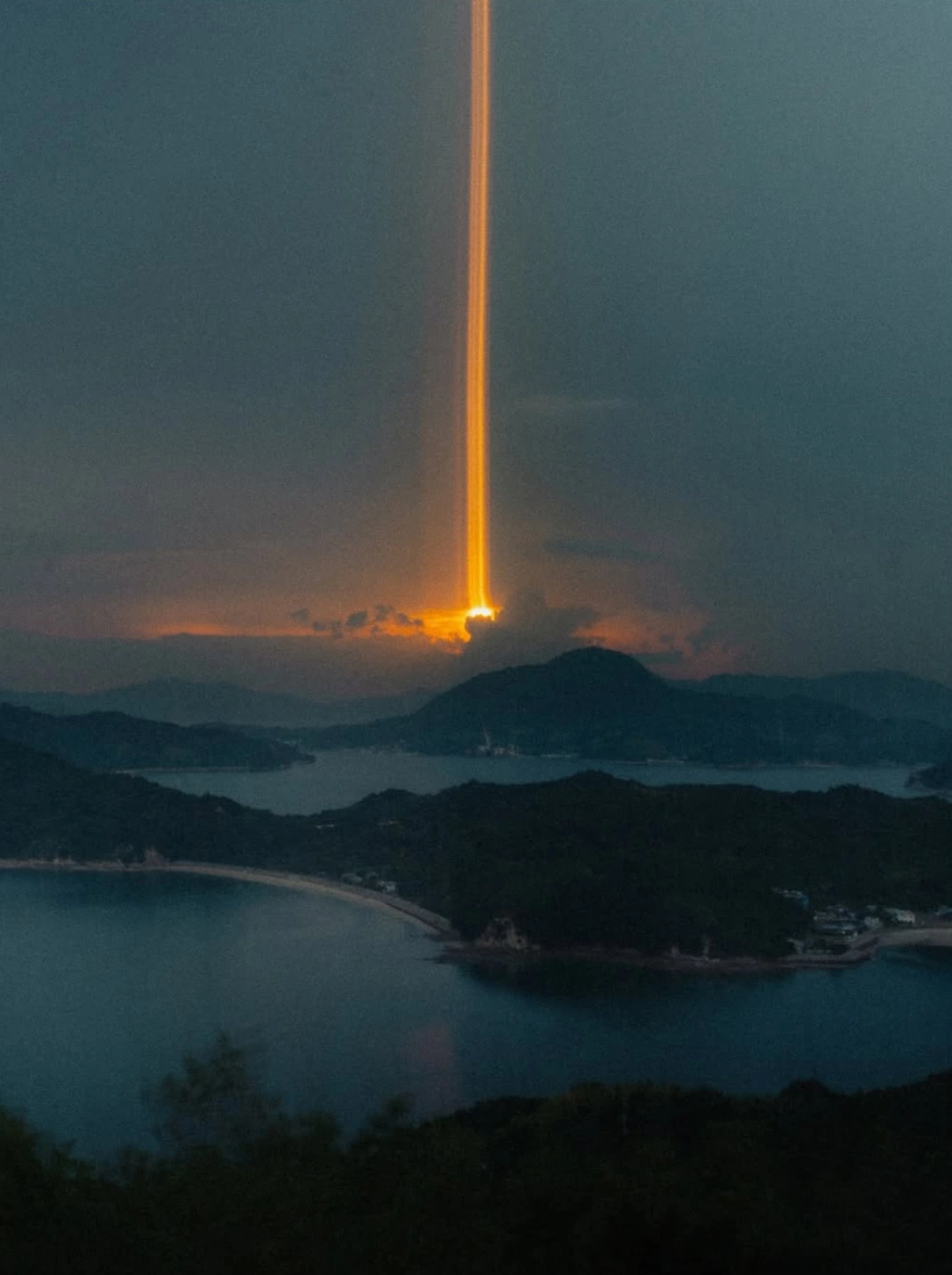 A bright orange beam of light breaking through clouds over water and mountains.
